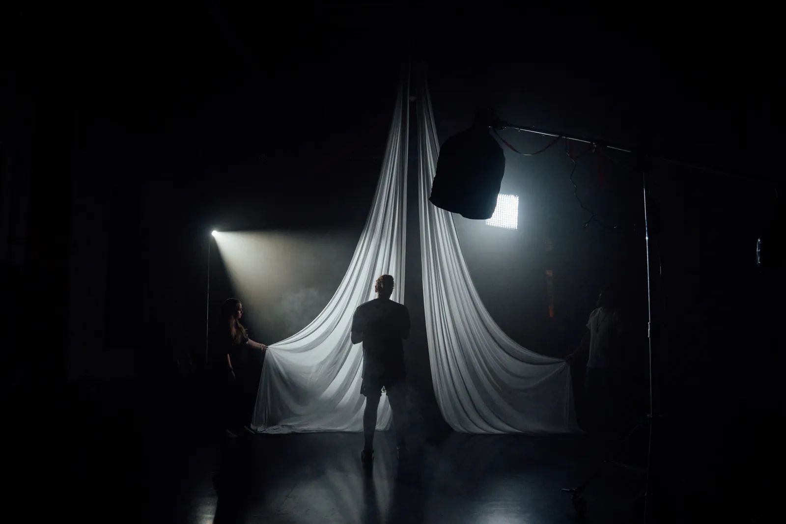 Behind-the-scenes photo of a dark set with hanging white silks, a haze-lit spotlight and crew holding the fabric.