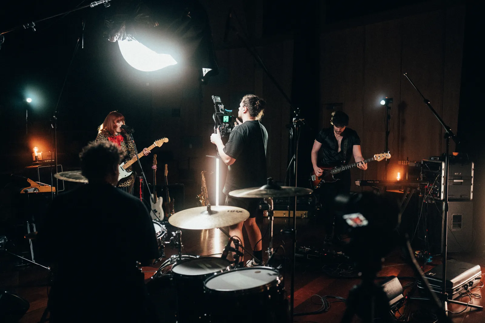 Behind-the-scenes photo of a band live session shot from behind the drum kit, with a camera operator filming the guitarist.