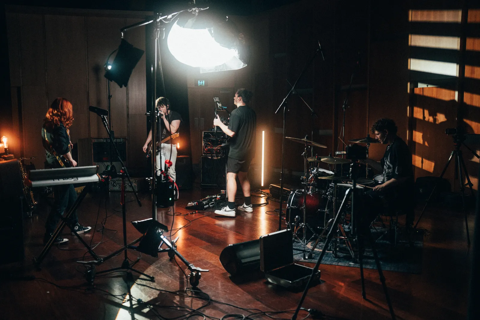 Behind-the-scenes photo of a full band live session set with a large softbox, practical lamps and a camera operator on the floor.