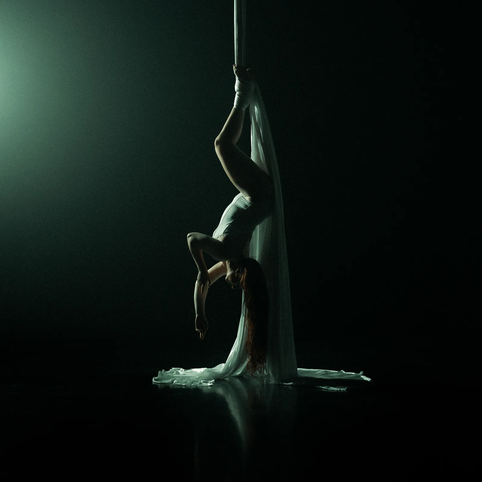 Aerial performer hanging upside down from white silks in a dark, green-lit space.